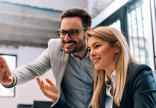 Un hombre y una mujer con ropa de negocios sonriendo y colaborando frente a una pantalla en una oficina